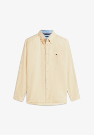 Beige pitkähihaiset napilliset paita, jossa on napitettu kaulus ja pieni logo vasemmassa rinnassa valkoisella taustalla.