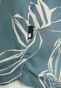 Gros plan d'un tissu bleu sarcelle avec un imprimé floral abstrait blanc et une petite étiquette rectangulaire noire et blanche cousue dans une couture.