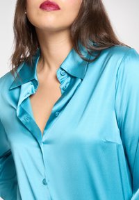 Turkusowa satynowa bluzka z klasycznym kołnierzem, guzikami z przodu i gładką fakturą. Posiada subtelną kieszeń po lewej stronie.