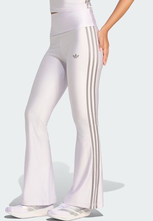 Person trägt weiße ausgestellte Leggings und passende Sneakers mit grauen Adidas-Streifen und Logo und steht vor einem einfarbigen hellen Hintergrund.