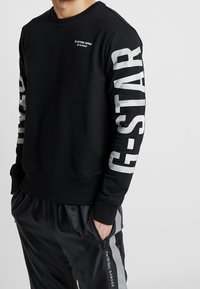 Černá mikina s bílým brandingem G-Star Raw na rukávech. Má kulatý výstřih a žebrované manžety. Nošena s černými tepláky.