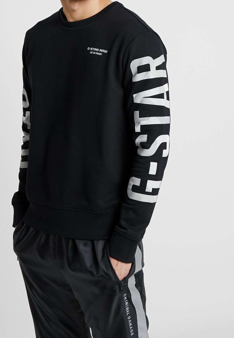 Černá mikina s bílým brandingem G-Star Raw na rukávech. Má kulatý výstřih a žebrované manžety. Nošena s černými tepláky.