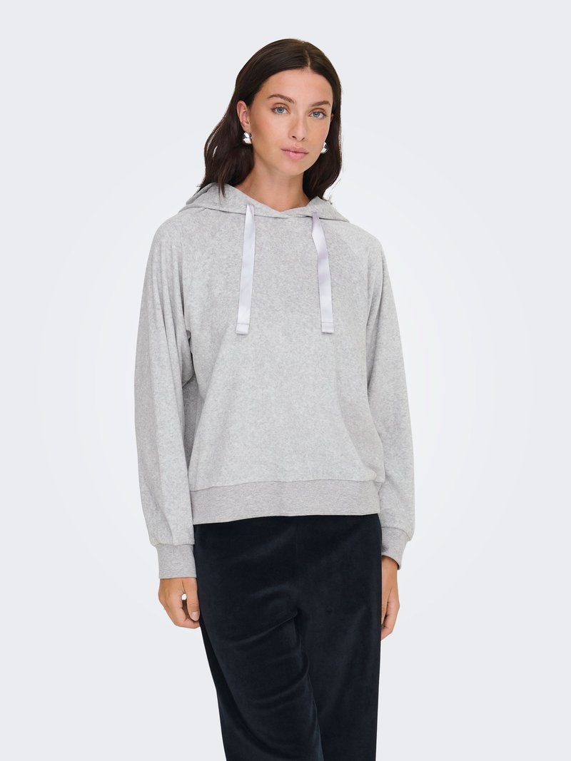 Femme aux cheveux foncés mi-longs, portant des boucles d'oreilles argentées, un sweat à capuche gris clair avec des cordons et un pantalon noir, debout devant un fond uni.