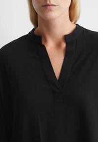 Tunic nera con scollatura a V e un piccolo dettaglio a taglio sul collo. Tessuto morbido, maniche lunghe e vestibilità comoda.
