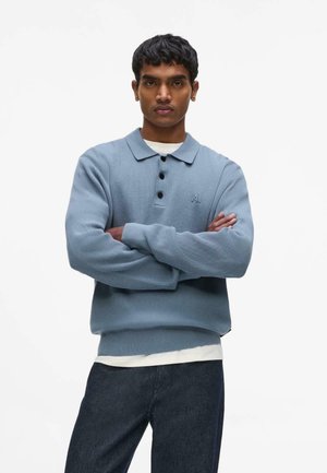 Junger Mann mit verschränkten Armen, der einen hellblauen gestrickten Polopullover über einem weißen Hemd und dunklen Jeans trägt, schaut frontal vor weißem Hintergrund.