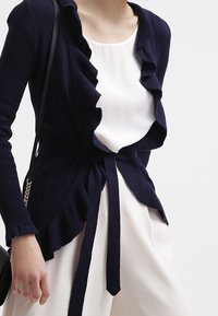 Námořnický žebrovaný kardigan s volánkovými lemy a zavazováním v pase, nošený přes bílý top, s hladkou texturou a přiléhavým střihem.