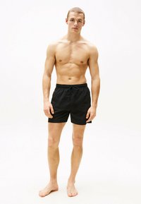 Pantaloni da nuoto neri con elastico in vita e stringa. Il modello è in piedi a piedi nudi con un fisico tonico, che mette in risalto la vestibilità dei pantaloni.