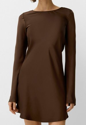Kobieta w długiej, ciemnobrązowej sukience za kolano z długim rękawem i dekoltem łódkowym, stojąca na jednolitym jasnym tle.