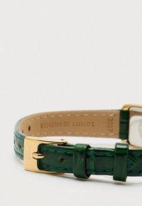 Green leather watch strap with glossy finish, gold-tone hardware, and white watch face. Features contrast stitching and a square buckle.