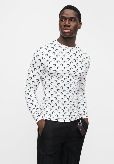 Man wearing fitted white long-sleeve shirt with black crescent pattern and black pants, hands in pockets, looking right on plain background.