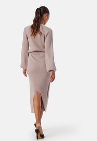 Brede midi-jurk in lichtbeige met ribbelstructuur, lange mouwen en een zijsplit, gecombineerd met zwarte hakken.