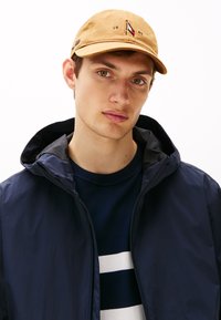 Beige caps med logo detalj, marineblå zip-up jakke med hette, og stripete marineblå, hvit og blå strikkegenser. Nærbilde av modellens overkropp.