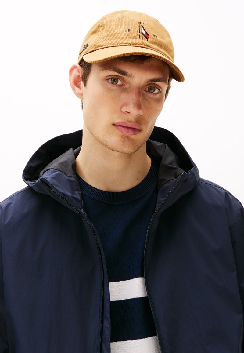 Beige caps med logo detalj, marineblå zip-up jakke med hette, og stripete marineblå, hvit og blå strikkegenser. Nærbilde av modellens overkropp.