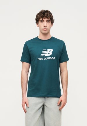 Mladý muž v tyrkysovom tričku s logom New Balance a svetlosivých nohaviciach, stojaci pred jednoduchým svetlým pozadím.