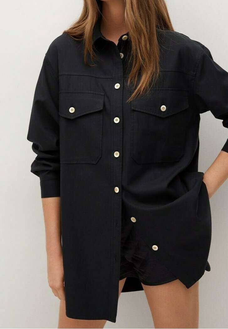 Femme portant une chemise noire oversize à boutons avec poches poitrine et boutons crème, associée à un short noir, une main dans la poche.