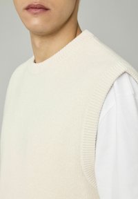 Pull en tricot sans manches crème avec des bords côtelés, superposé sur une chemise blanche à manches longues. Texture lisse avec un col rond.
