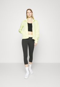 Completo sportivo con una giacca leggera gialla zip, un top nero corto e leggings capri neri con un motivo laterale, abbinato a sneakers bianche.