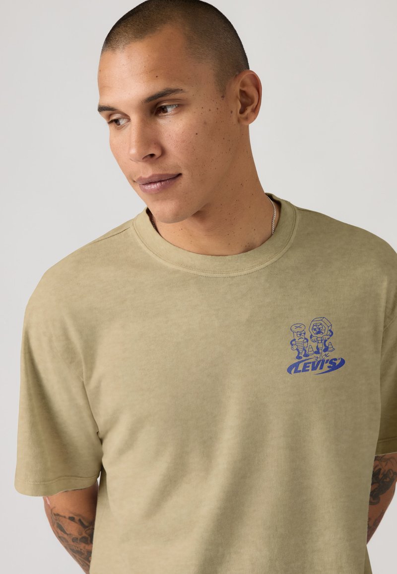 Mann mit kurzen Haaren trägt ein beiges Levi's-T-Shirt mit einem kleinen blauen Motiv von zwei Cartoon-Figuren auf der Brust und schaut nach links.