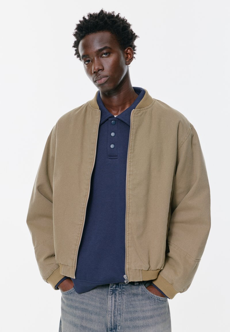 Giacca bomber color beige con polsini e colletto a costine, chiusura con zip, indossata sopra una polo blu navy, abbinata a jeans di denim chiaro. Sfondo semplice.
