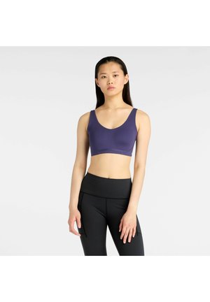 Vrouw draagt een paarse sportbh en zwarte legging met hoge taille, staand met een neutrale gezichtsuitdrukking tegen een effen lichte achtergrond.