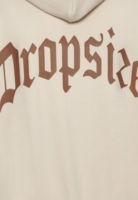 Καφέ γοτθικό κείμενο «Dropside» σε κρεμ ύφασμα με μέρος κουκούλας ορατό στο πάνω μέρος του ενδύματος.