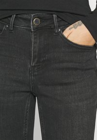 Μαύρα τζιν με κλασικό σχέδιο πέντε τσέπες, με χρυσό κουμπί και διακριτές ραφές, εκτεθειμένα σε ουδέτερο φόντο.