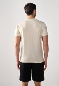 Beige kortermet t-skjorte laget av mykt materiale, med et enkelt design. Båret med mørke shorts, viser en ren, rettkuttet bakside.