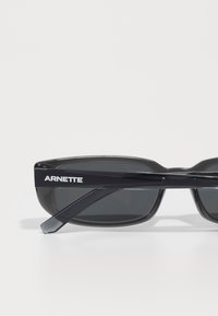 Lunettes de soleil noires avec monture rectangulaire. Présentent des verres sombres, un matériau plastique brillant et une marque "ARNETTE" en blanc sur la branche.