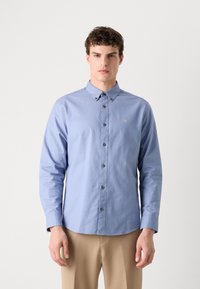 Camisa azul clara de botão, com colarinho pontudo, mangas longas e botões escuros. Apresenta um pequeno logotipo dourado no peito.