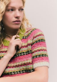Kötött pulóver több színű csíkokkal rózsaszín, zöld és fekete színekben, rövid ujjakkal és kontrasztos zöld gallérral.