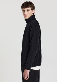 Modello maschile in un maglione nero a collo alto e pantaloni neri, in piedi di profilo contro uno sfondo chiaro e semplice.