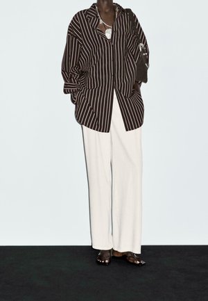 Person wearing oversized black and white vertical striped shirt, white wide-leg pants, black sandals, silver necklace, and holding a black handbag.