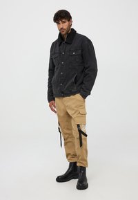 Giacca in denim nero con colletto in sherpa, pantaloni cargo beige con tasche e cinture, e stivali neri alla caviglia con suola spessa.