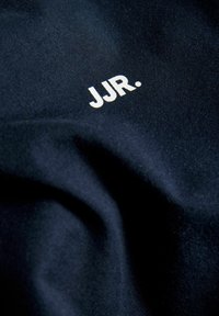 Σκούρο μπλε ύφασμα με λευκό κείμενο "JJR." τυπωμένο κοντά στο κέντρο, δείχνοντας υφή και ελαφριές πτυχώσεις στο υλικό.