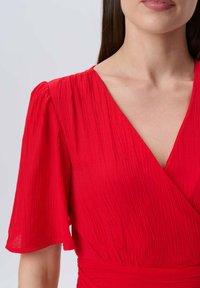 Rotes Kleid mit kurzen, luftigen Ärmeln, ausgestattet mit einem tiefen V-Ausschnitt und strukturiertem Stoff. Die Taille ist tailliert, was die Silhouette betont.