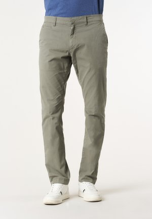 Uomo che indossa pantaloni slim verde oliva e sneakers bianche, in piedi contro uno sfondo semplice.