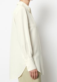 Lange mouwen, crèmekleurig shirt met een klassieke kraag, licht getailleerde pasvorm en knoopmanchetten. De stof lijkt glad en lichtgewicht te zijn.