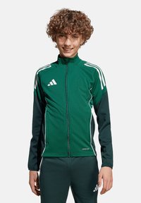 Groene trainingsjack met een hoge kraag, volledige rits, witte accenten en drie witte strepen op elke schouder. Het materiaal lijkt glad en sportief.