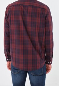 Una persona che indossa una camicia a quadri rossa e blu navy e jeans blu, vista di spalle, con maniche corte allacciate ai polsi.