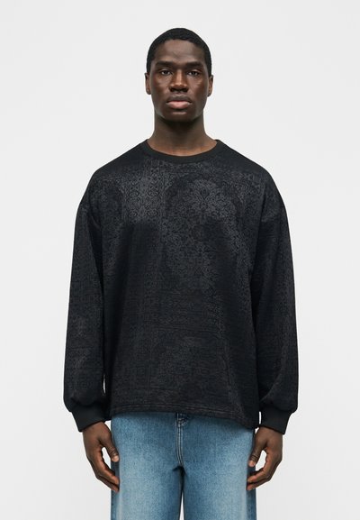 Άνδρας που στέκεται και κοιτάζει μπροστά, φοράει μακρυμάνικο μαύρο πουκάμισο με μοτίβα και ανοιχτόχρωμα μπλε τζιν, με λευκό απλό φόντο.