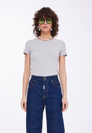 T-shirt grigio a maniche corte abbinato a jeans blu in denim a vita alta. Gli accessori includono occhiali da sole verdi e collane di perle sovrapposte.