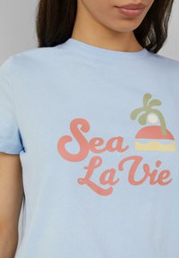 Lichtblauw katoen t-shirt met een kleurrijk bedrukt ontwerp met de tekst "Sea La Vie" en een palmboom graphic. Korte mouwen, standaard pasvorm.