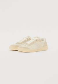 Lys beige sneakers med teksturert suede og glatt overdel, med hvite snøringer og en lett polstret krage, på en teksturert gummisåle.
