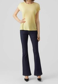 Žena v žlutém krátkém rukávu, tmavě modrých rozšířených džínách a černých botách na vysokém podpatku, stojící s jednou rukou na boku.