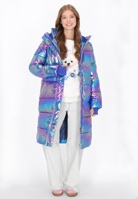 Irideszcens hosszú pufajka kék és lila árnyalatokban, cipzáras záródással, kapucnival és rugalmas mandzsettákkal; egy fehér pulóver és világos farmernadrág felett viselve.