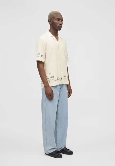 Άνδρας με κοντά ξανθά μαλλιά που φοράει κρεμ πουκάμισο με κοντά μανίκια και κουμπιά με μαύρα σχέδια από φοίνικες και ανοιχτό μπλε τζιν παντελόνι με φαρδιά πόδια.