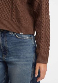 Bruine gebreide trui met kabelpatroon, geribbelde manchetten, gecombineerd met blauwe denim jeans met een voorzak en gebogen naaddetails.
