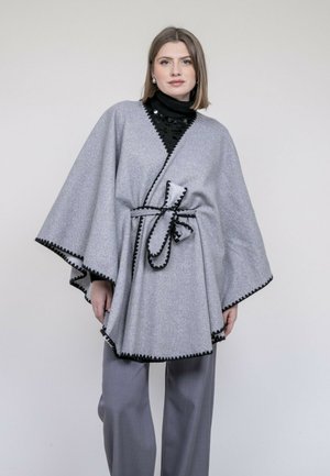 Femme debout dans un poncho gris avec bordure et ceinture noires, col roulé noir et pantalon gris, sur un fond clair et uni.