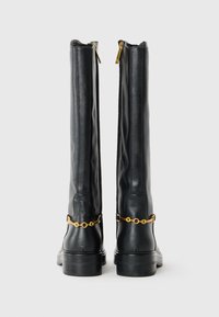 Bottes en cuir noir hautes avec un détail de chaîne dorée à l'arrière et des tirettes de fermeture éclair dorées, se tenant côte à côte sur un fond blanc.