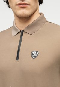 Μπεζ πόλο πουκάμισο με φερμουάρ στον γιακά, που φέρει μαύρο λογότυπο. Το ύφασμα έχει λεία υφή και διακριτική λάμψη.